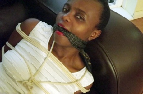 Black female is left tied and cleave gagged in a white dress on a leather sofa